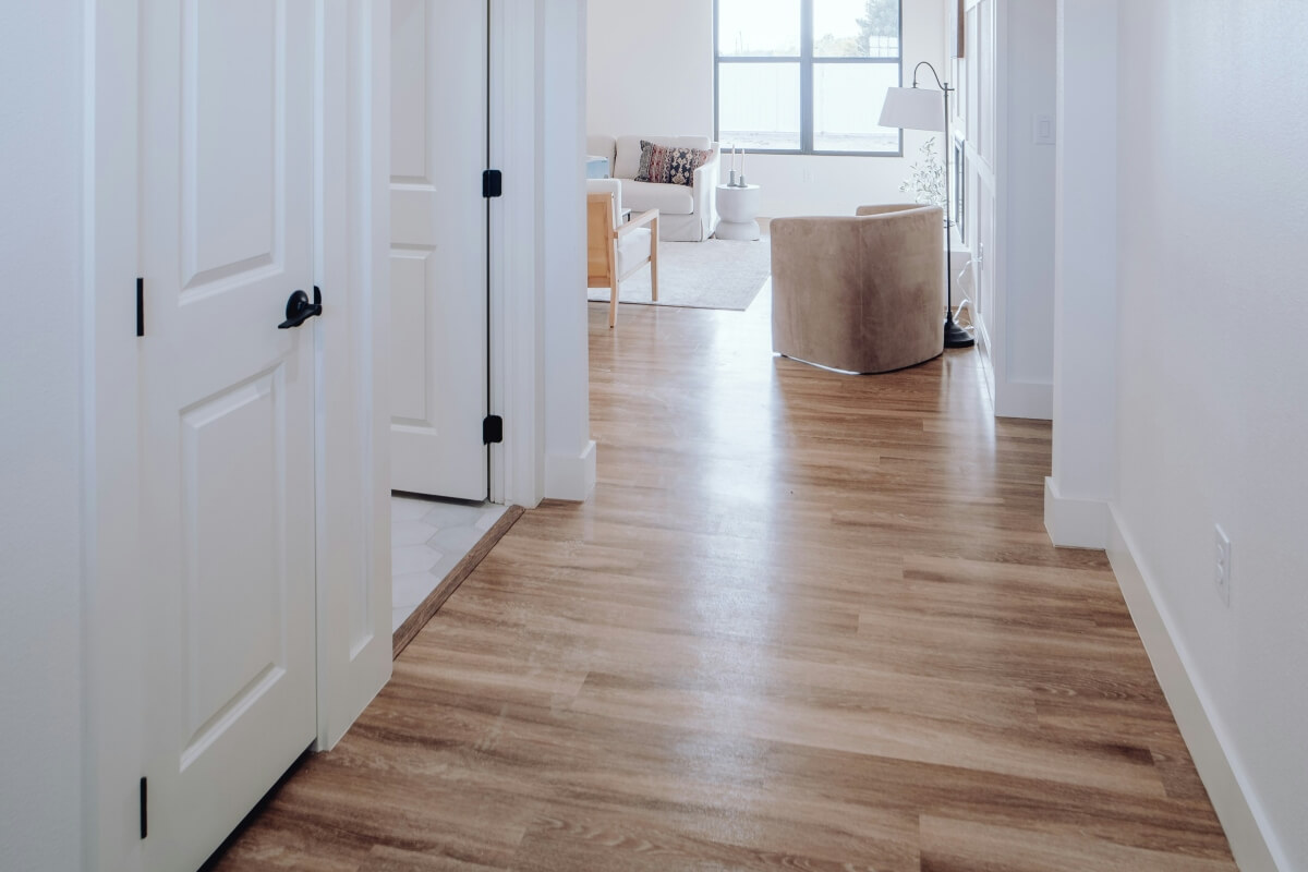 Bright hallway opening into a sunlit living room with white walls, wood flooring, and a beige armchair by a window.
