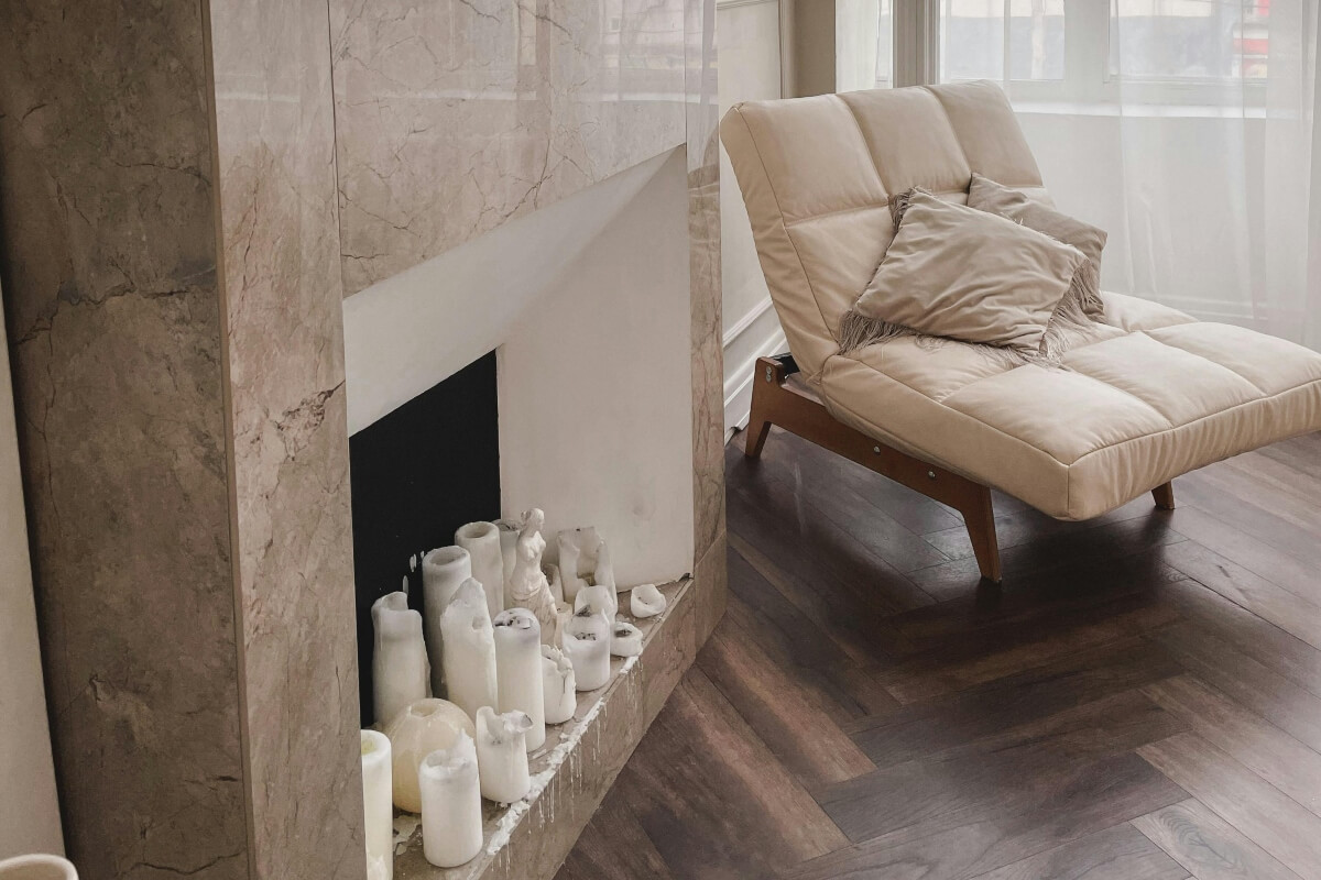 Beige chaise lounge with two pillows beside a marble fireplace and a row of white candles on the hearth in a living room corner.