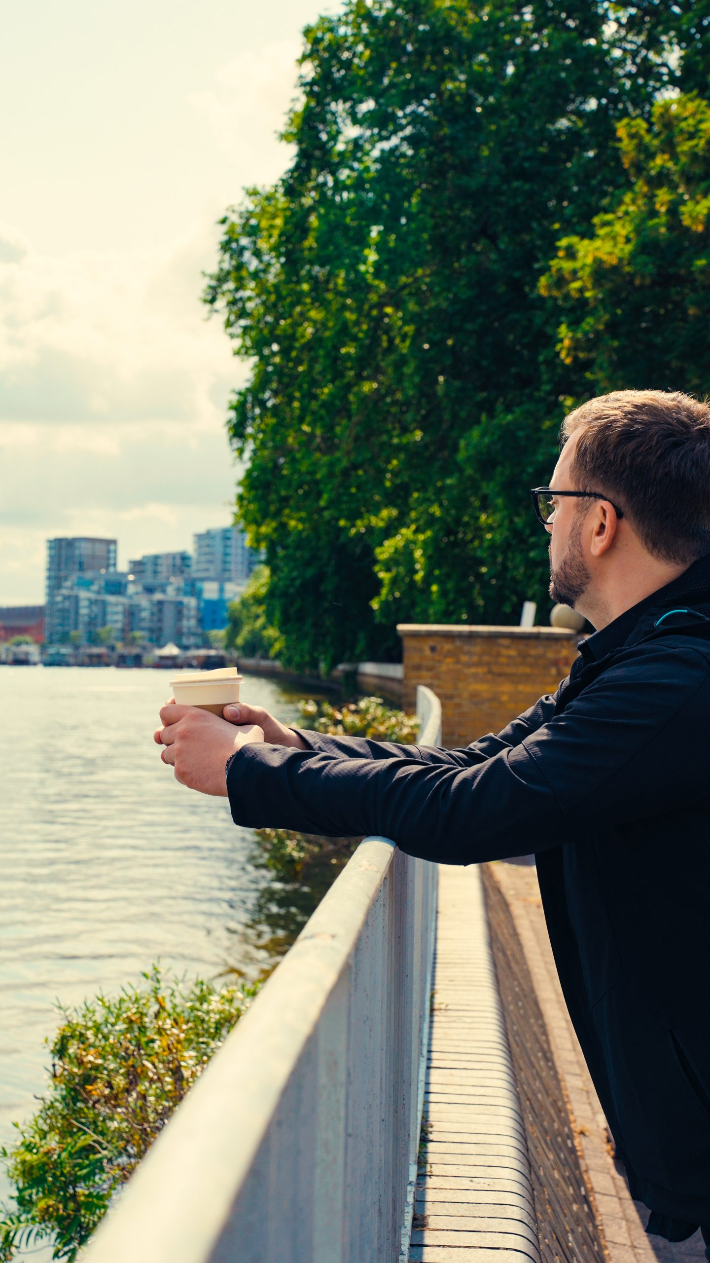 Marcin Kluk leaning on a railing by the river, holding a cup of coffee, with trees and city buildings in the background.