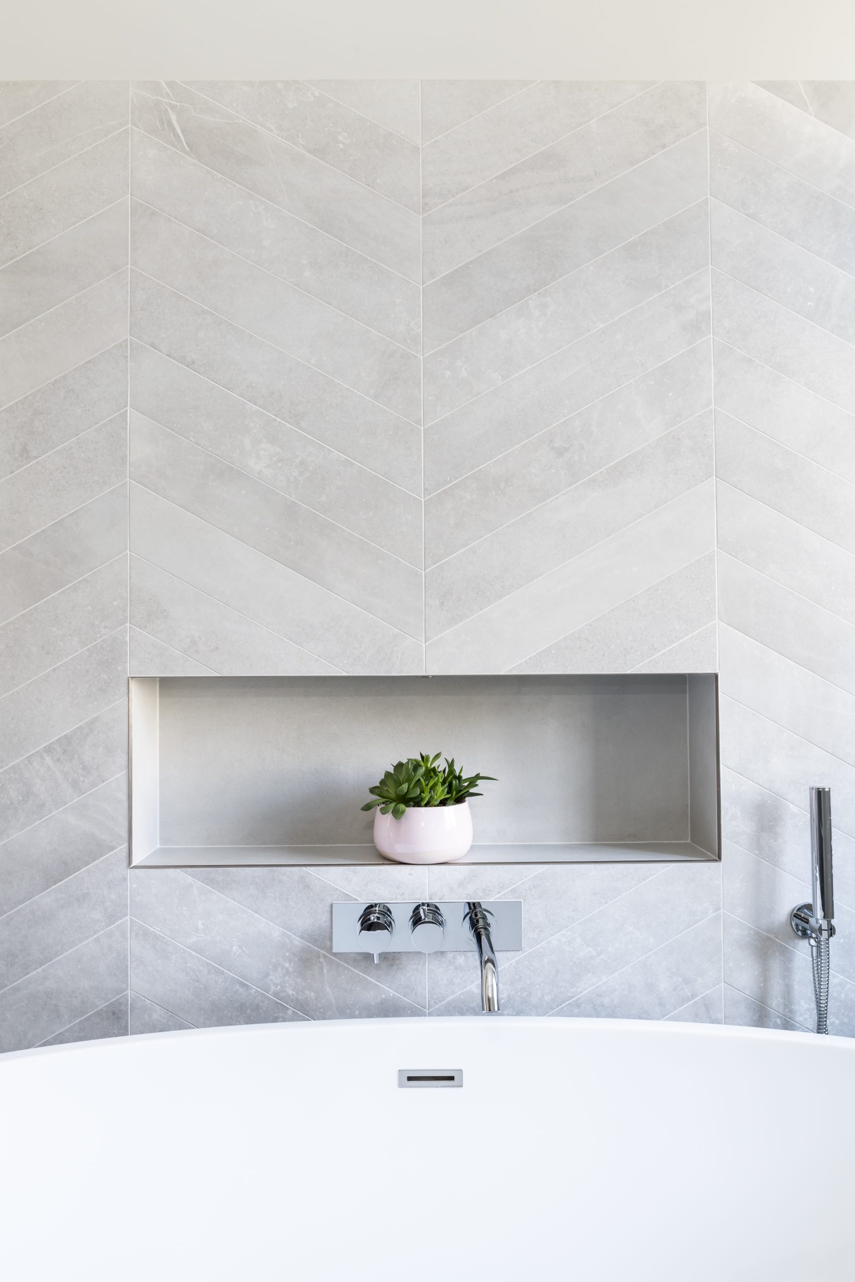 Freestanding bath with recessed wall niche, chevron porcelain tiles, and concealed chrome bath filler.