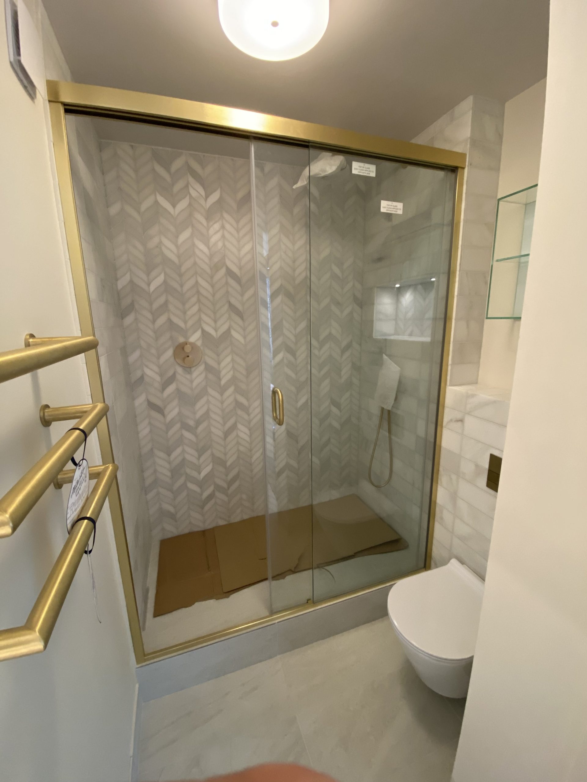 Gold-framed glass shower with chevron tile walls, handheld shower, and cardboard mat on the shower floor; toilet visible to the right.