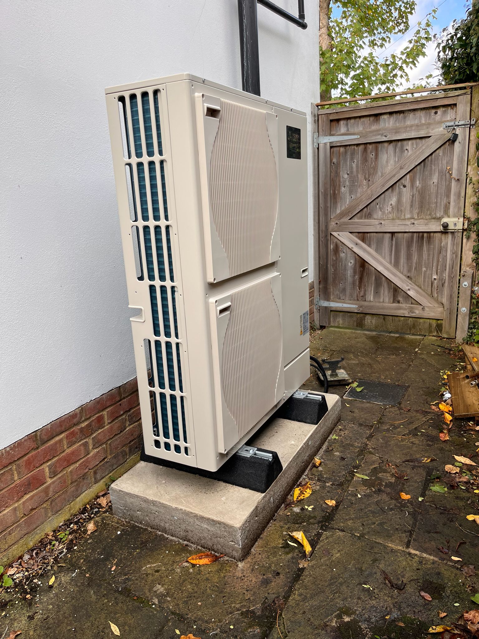 Outdoor air conditioning condenser unit on a concrete pad beside a white wall and wooden gate