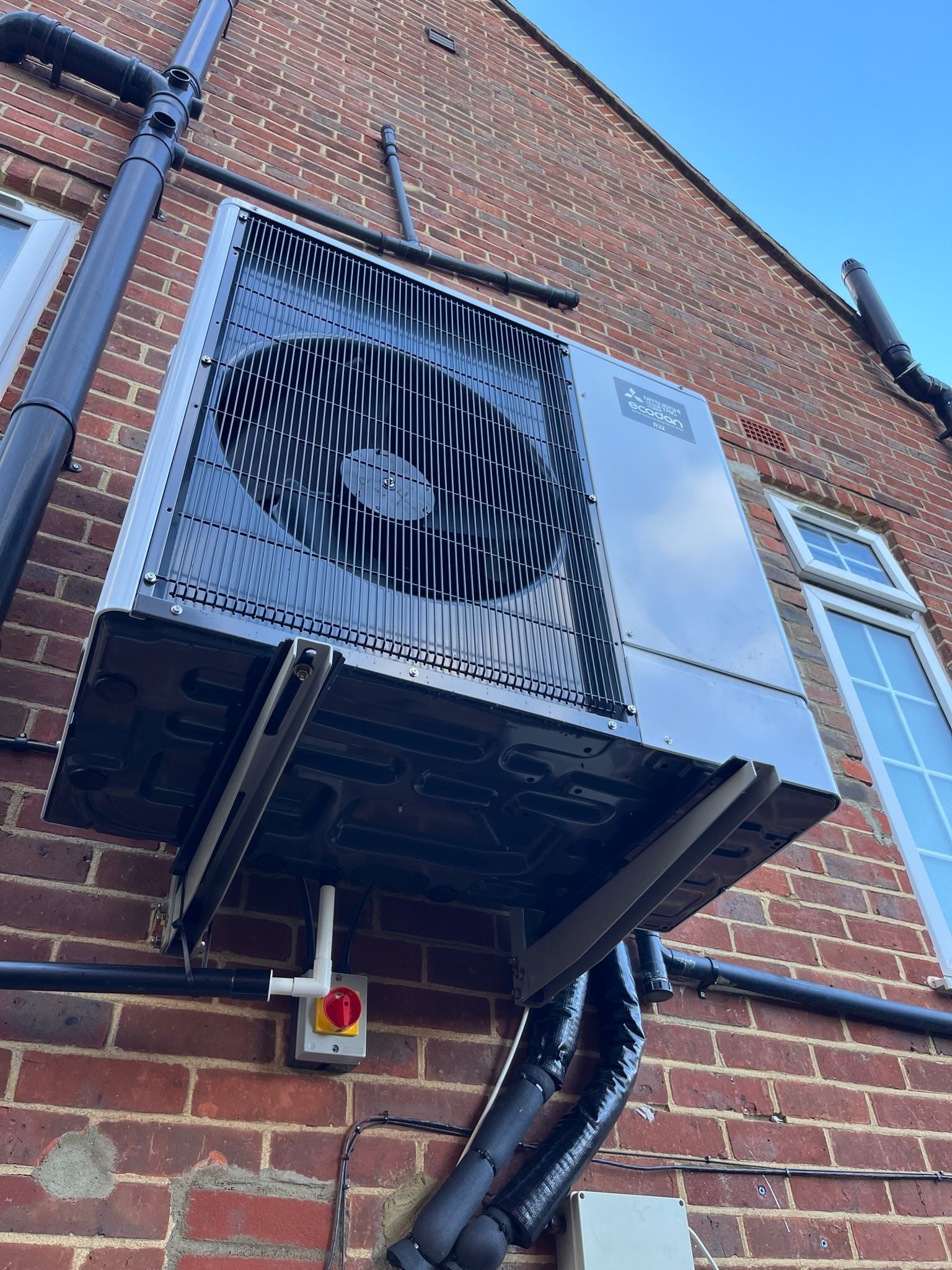Air source heat pump mounted on exterior brick wall of UK home with insulated pipework and electrical isolation switch.