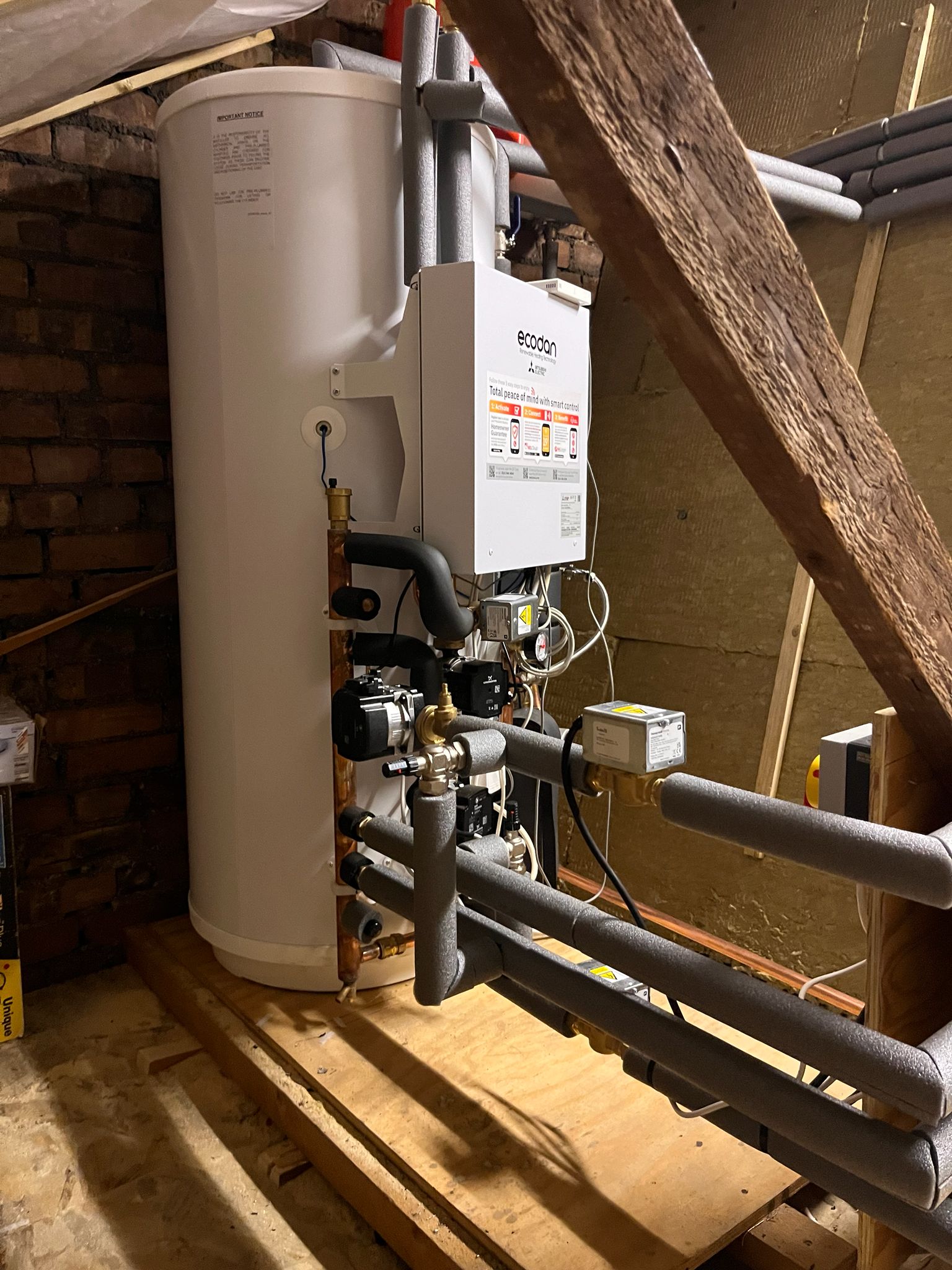 Heat pump hot water cylinder and hydraulic unit installed in loft space with insulated pipework and heating controls.