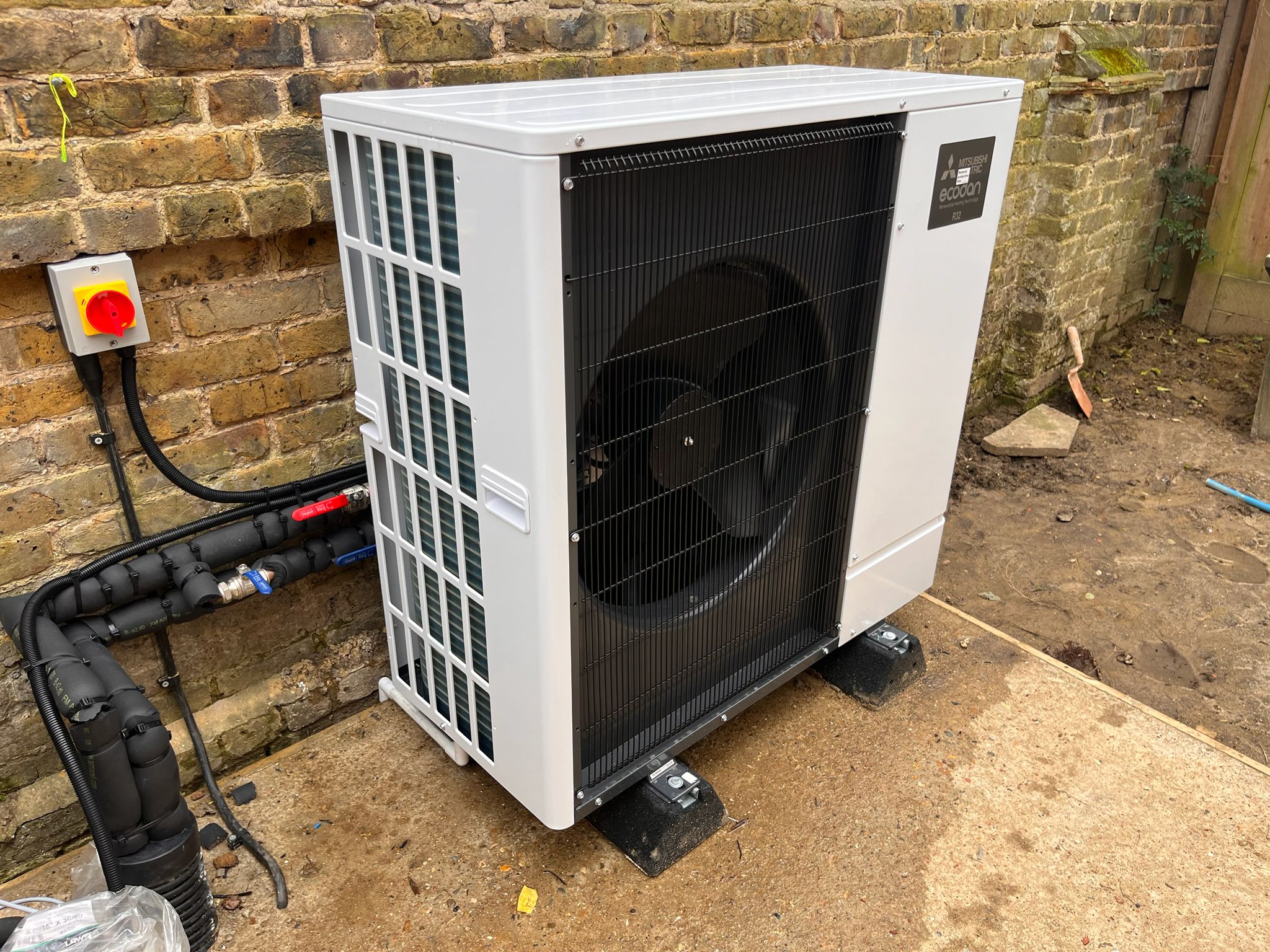 Outdoor Mitsubishi HVAC condenser unit mounted beside a brick wall, with pipes and a red/yellow electrical disconnect switch nearby. It’s an outdoor air conditioning unit.