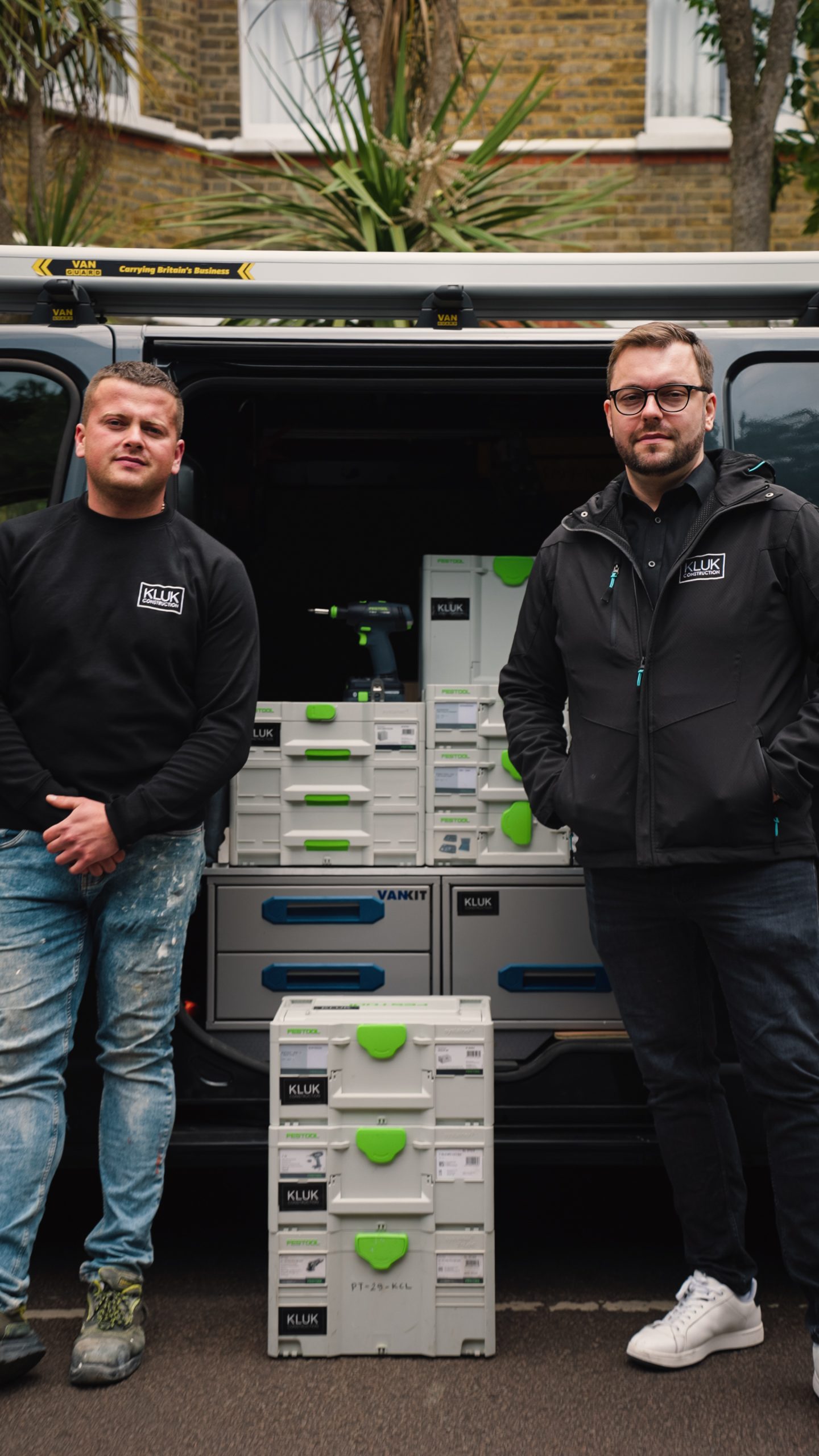 Two construction workers stand by an open van filled with stacked toolboxes and a drill set, KLUK branding visible on boxes and clothing.