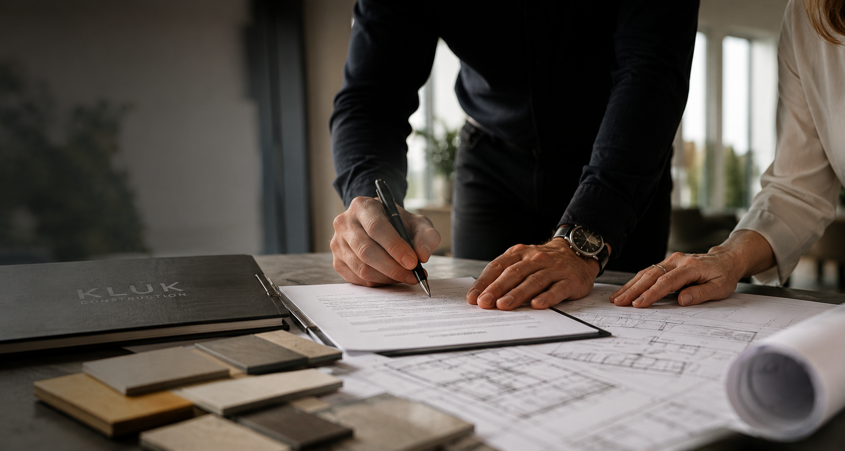 Two professionals sign a contract over architectural plans, with a KLUK Construction binder and color swatches on the table nearby.
